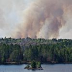 A dark smoke plume is visible above the forested island, lake in the foreground.