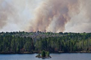 A dark smoke plume is visible above the forested island, lake in the foreground.