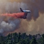 Heavy aircraft flying in dark smoke and dropping red retardant over the forest.