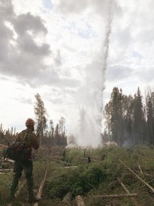 Wildland firefighter standing a an open forest and holding a handheld radio looks at an erupting geyser.