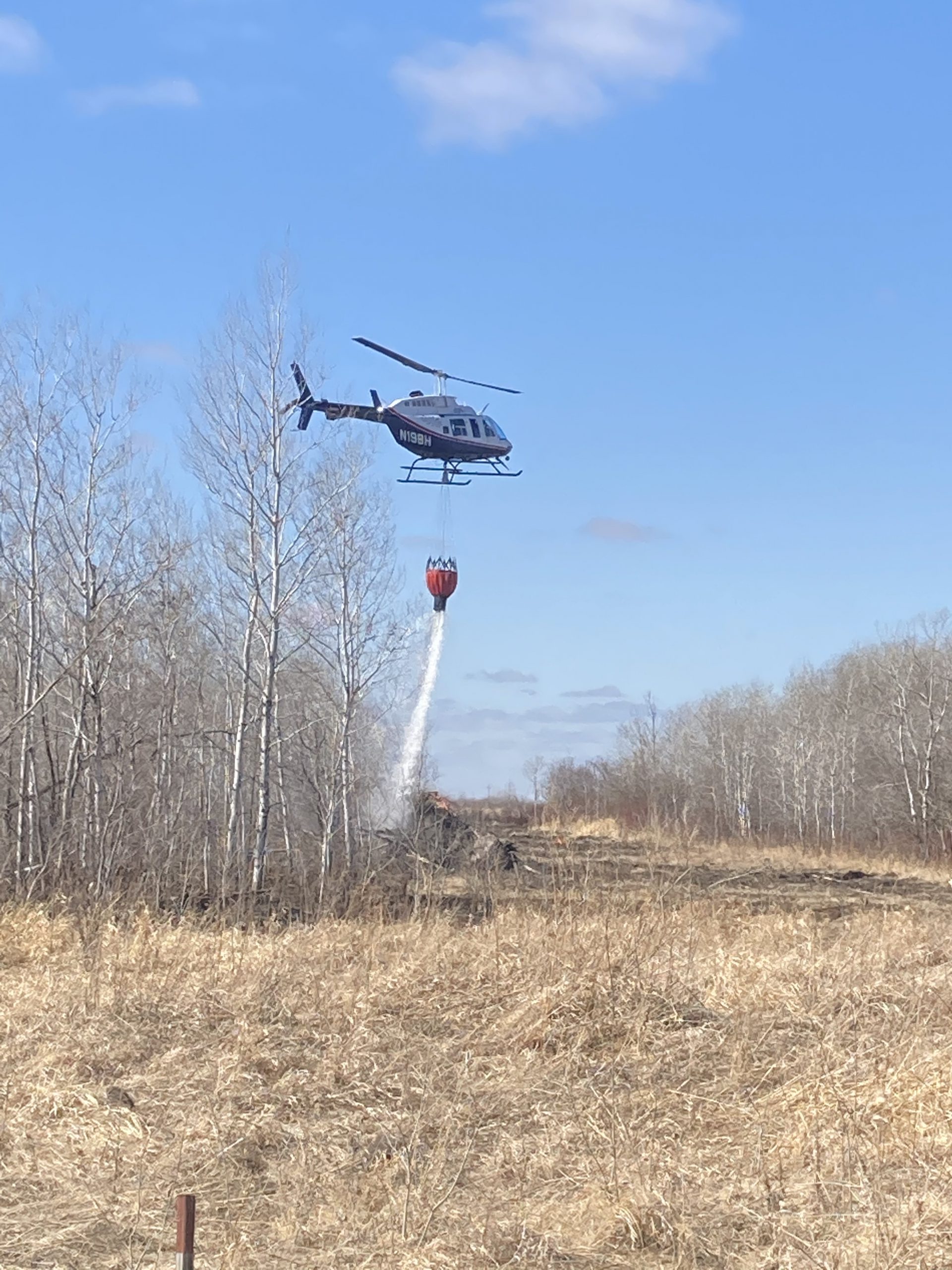 Helicopter releases water from bucket over a field and trees.