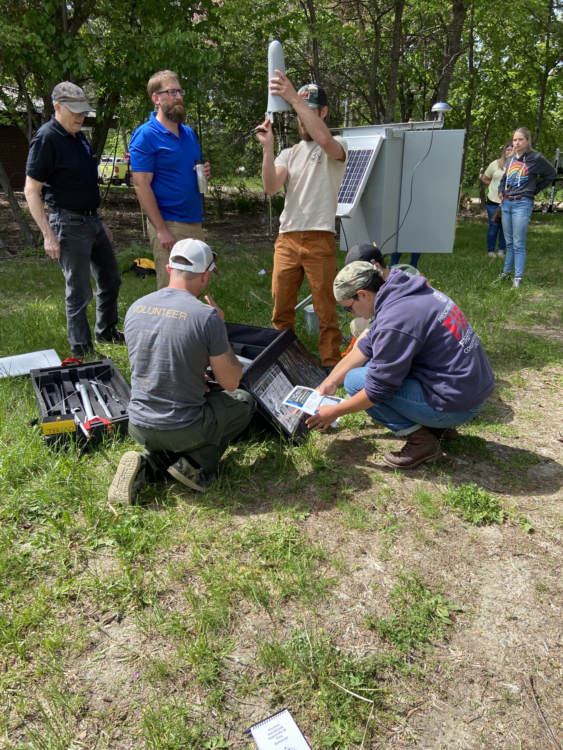 Eight people work to set up a weather station in a shaded area outside.