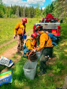 Three people in yellow shirts and red hardhats in front of a red fire engine work to fill up a bucket of water.