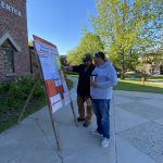 Two people stand and look at an information sandwich board outside of a brick building.