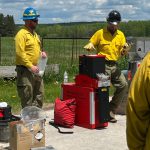 Two people in yellow shirts and wearing hard hats stand next to a small red machine.