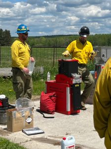 Two people in yellow shirts and wearing hard hats stand next to a small red machine.