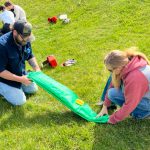 Two people sitting on the ground work on rolling up a green tarp.
