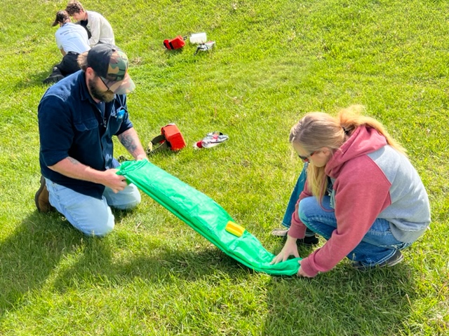Two people sitting on the ground work on rolling up a green tarp.
