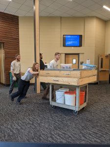 A person pushes a cart holding a wood box on top and buckets beneath the wood box, through a hallway.