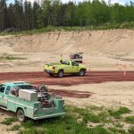 A green and yellow vehicle drive through a gravel pit with trees in the distance.