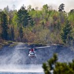 A helicopter drawing water from a lake surrounded by forest.