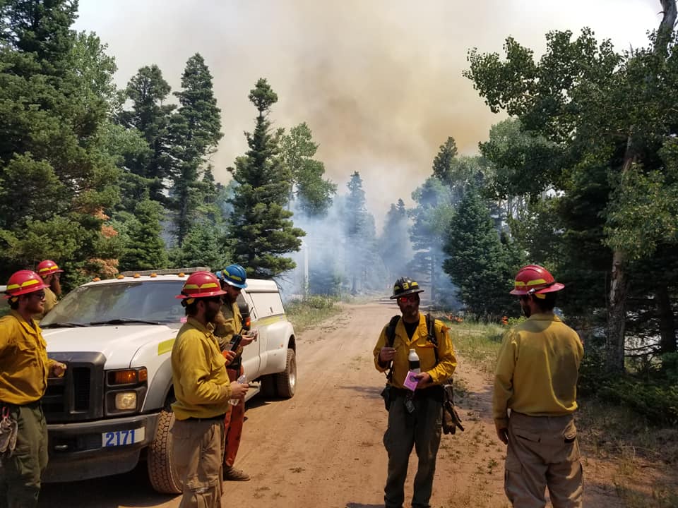 A group of five firefighters dressed in yellow shirts meet near a white truck on a gravel road.