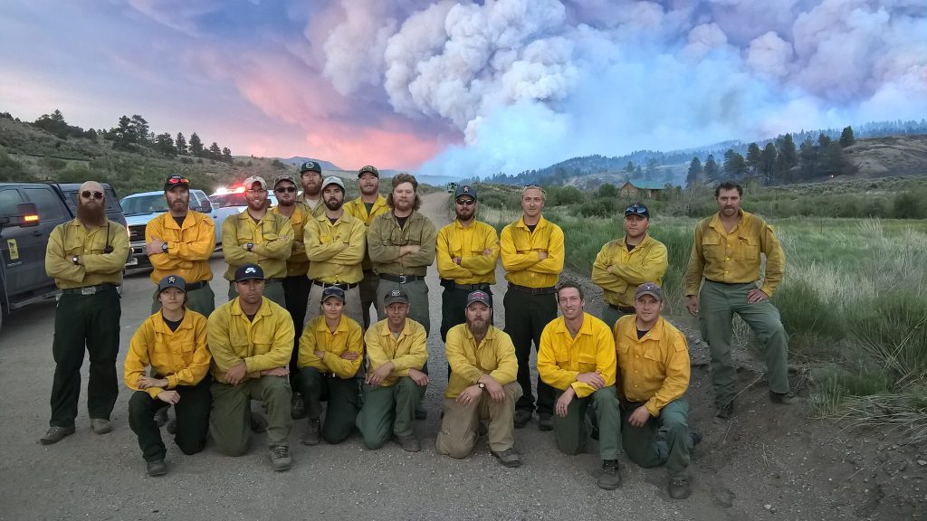A group of 19 people dressing yellow fire shirts pose for a group photo on a gravel road with bellowing smoke rising above the landscape in the background.