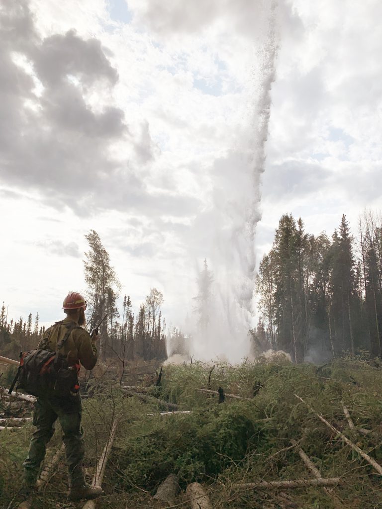 A wildland firefighter holding a radio and wearing a backpack looks toward a geyser bursting toward the sky.