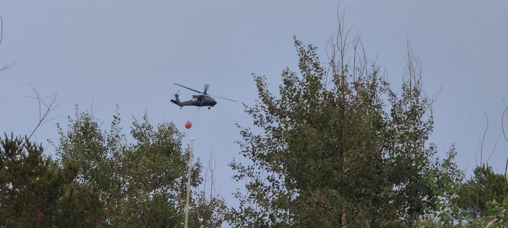 Black helicopter with bucket above trees.