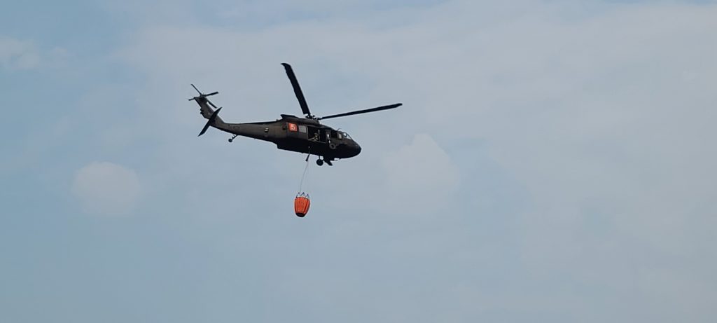 Black helicopter with orange bucket in the sky.