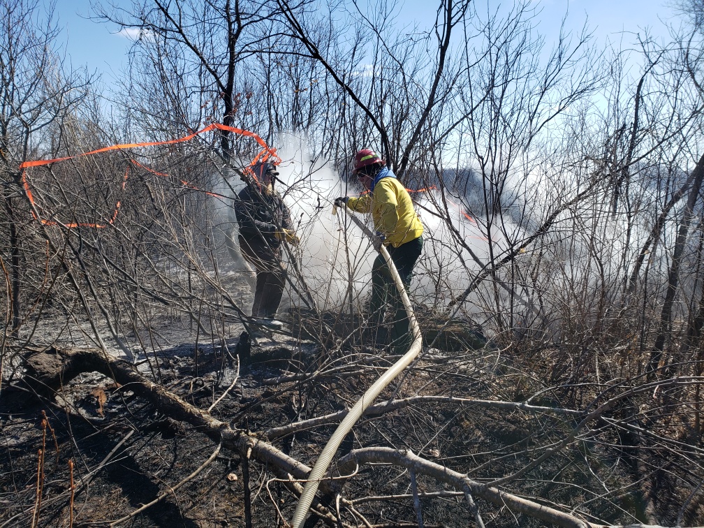 Two firefighters spray water on smoky brush with hose
