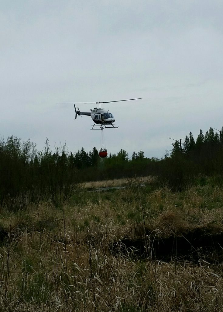 Helicopter with bucket above a marsh area.