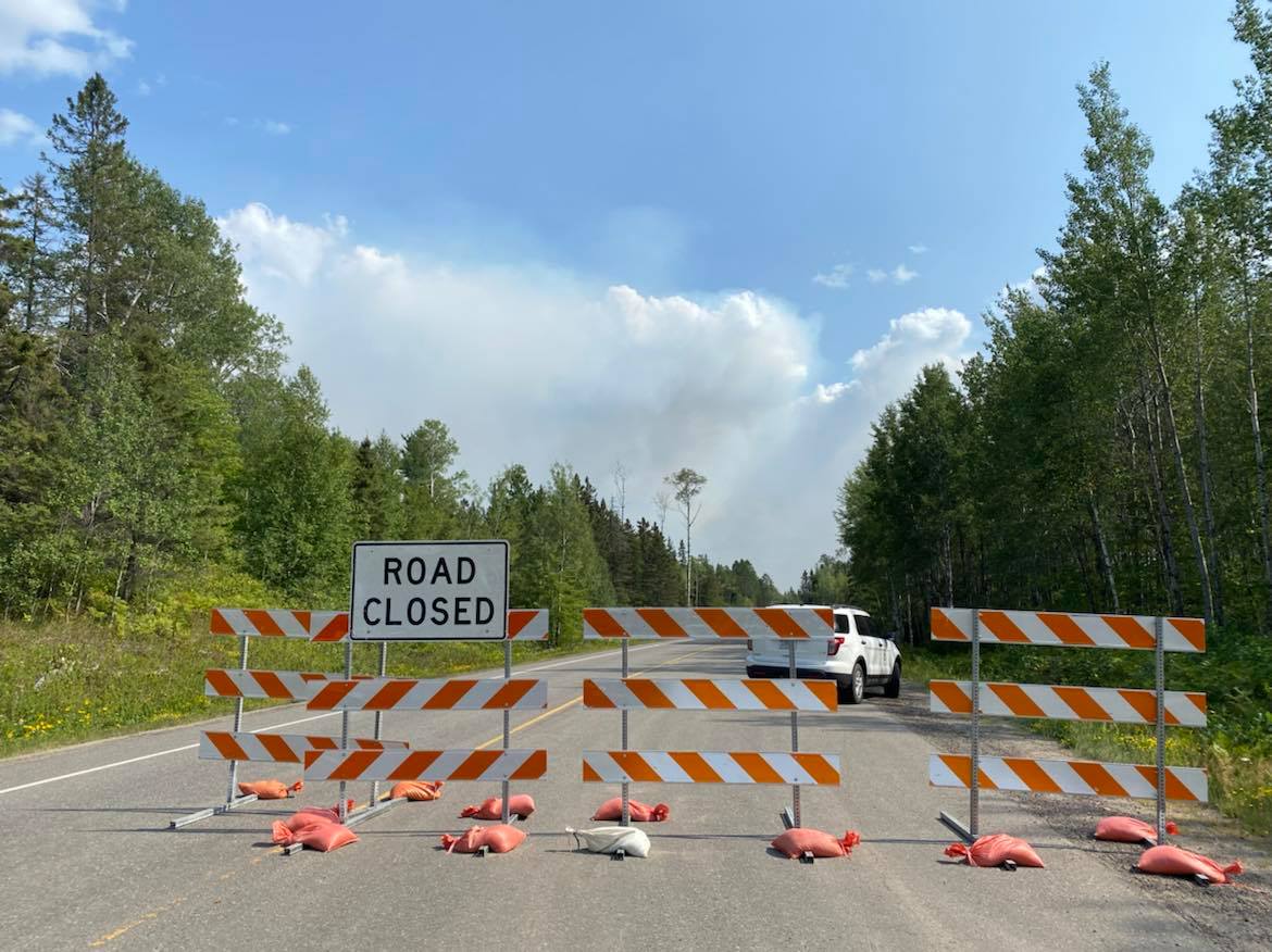 Road closure sign next to forest with smoke rising in blue sky..