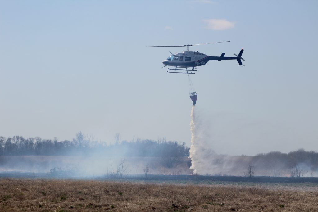 Helicopter releases water from bucket on to a field near trees.