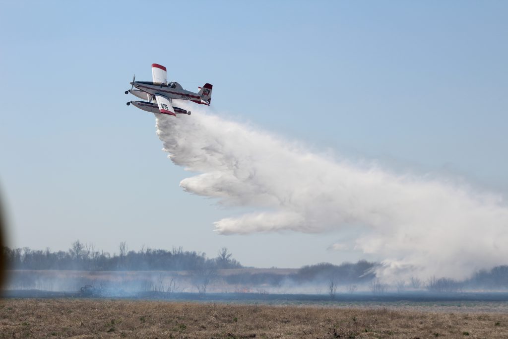 Fire boss aircraft releases water onto flames in a field.