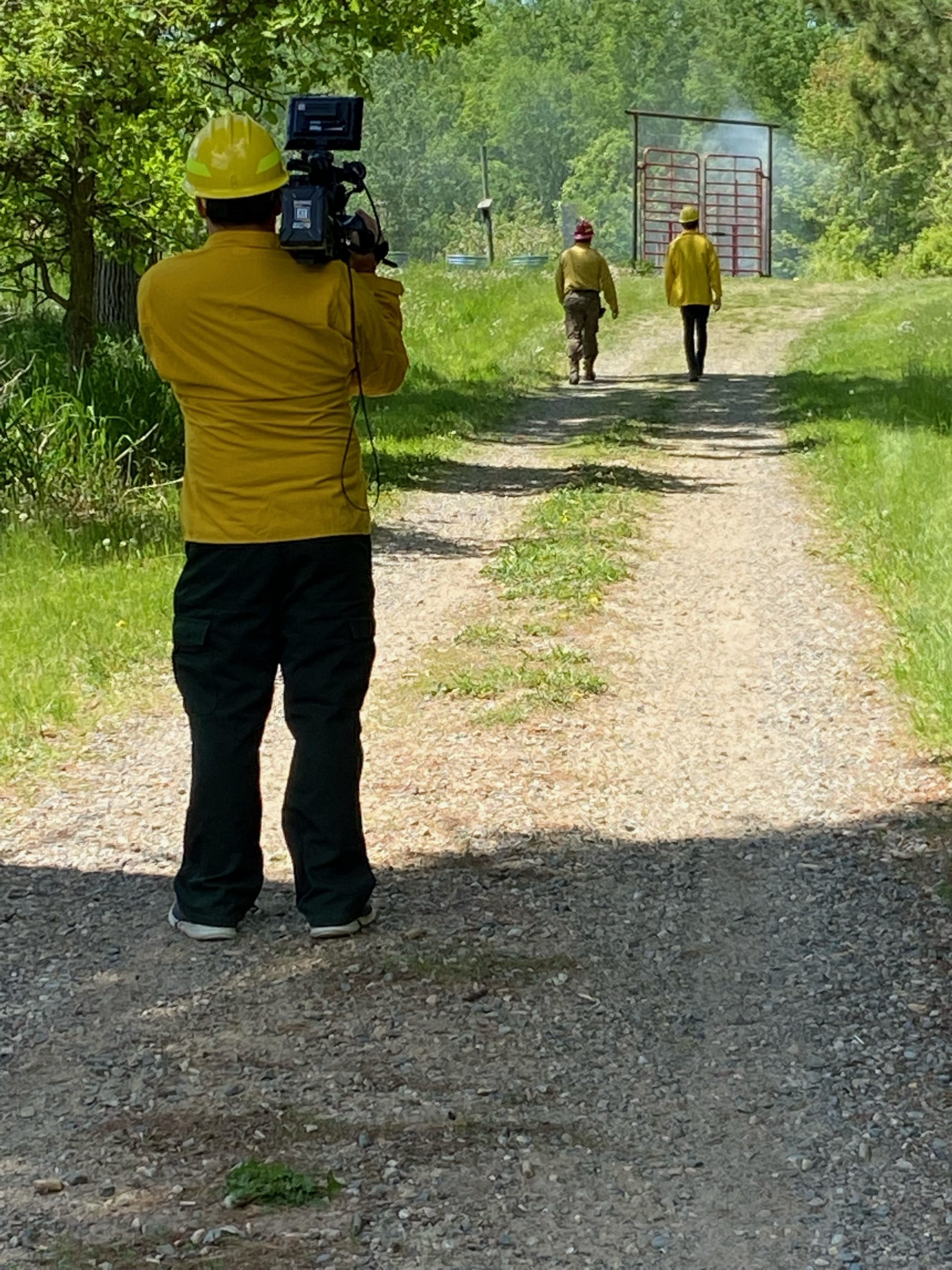 A person in a yellow shirt holding a large camera pointed at two people in yellow shirts walking down a dirt path.