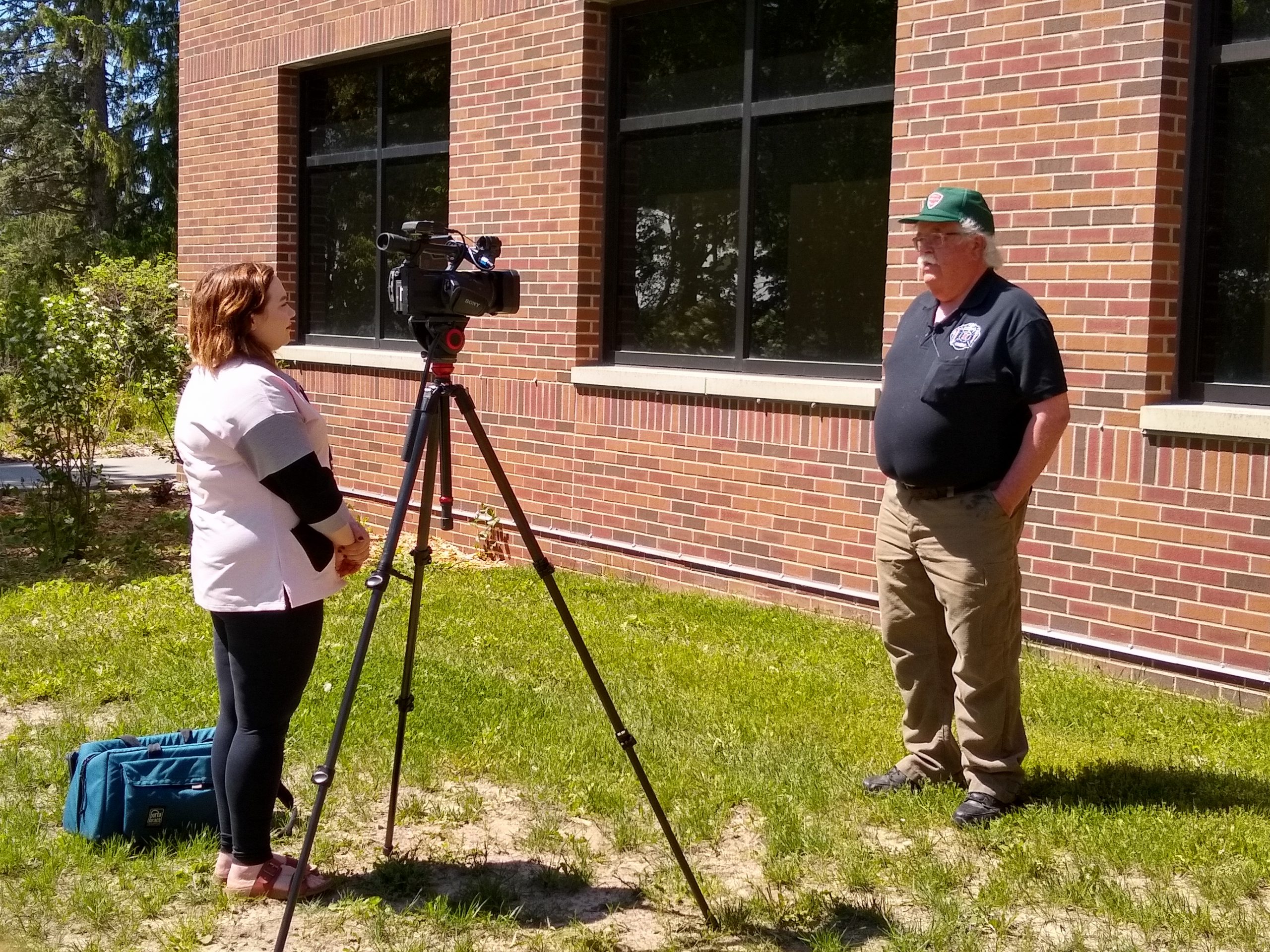 Reporter with a camer interviews a man in a dark shirt and green hat.