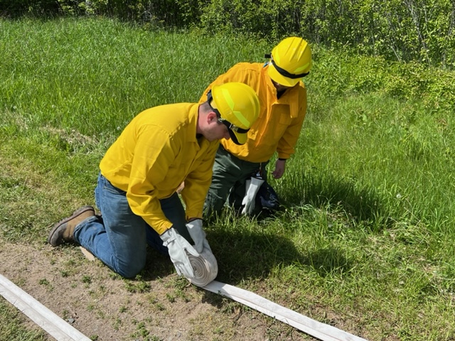 Two people in yellow shirts and yellow hardhats roll up a white firehose that's lying on green grass.
