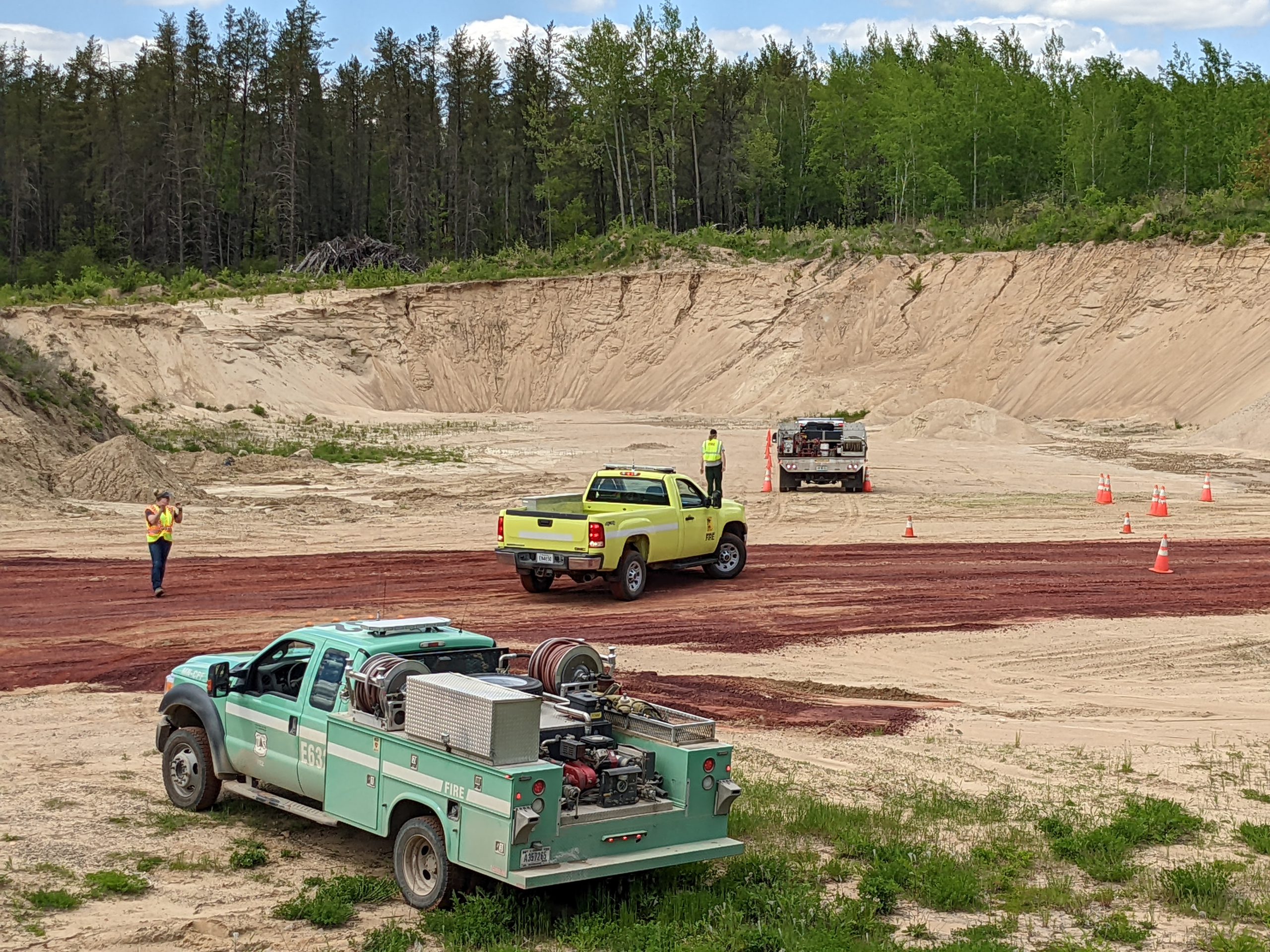 Green and yellow trucks in a gravel pit with people standing in the background.