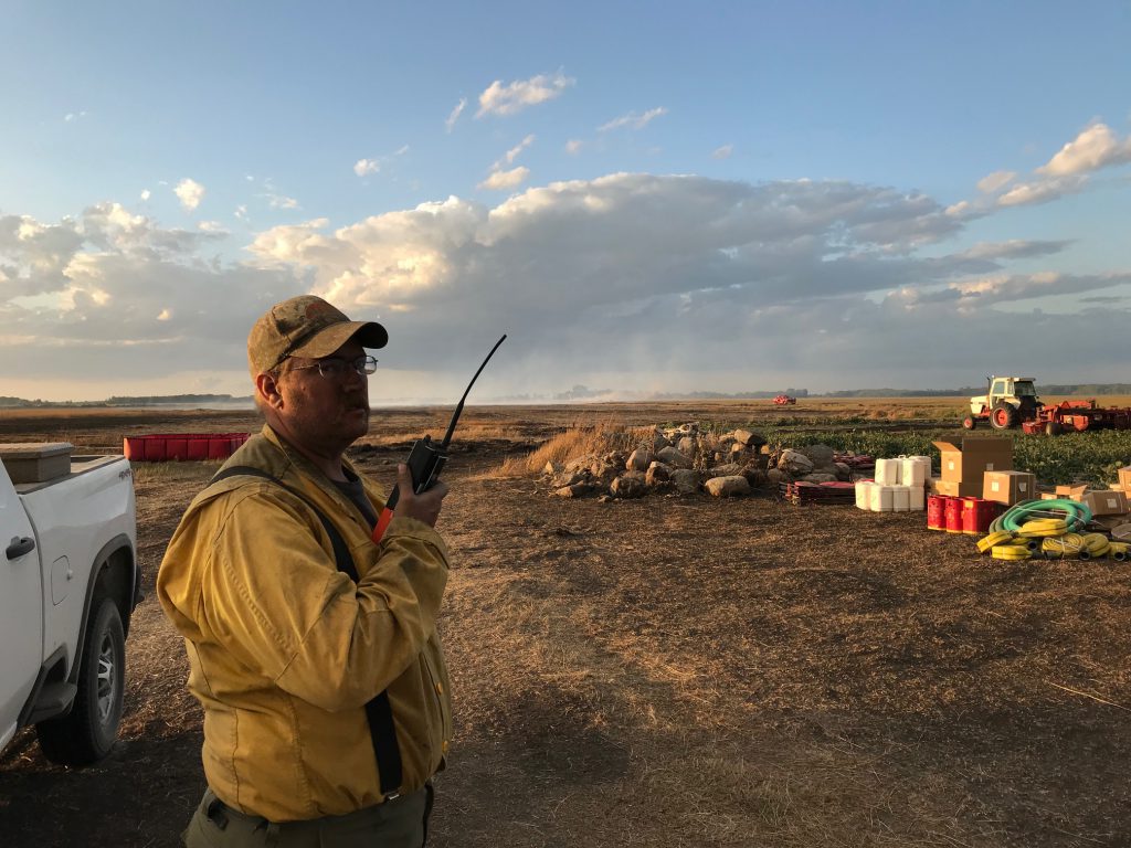 Mat in yellow shirt holding a radio stands in a dry open field.