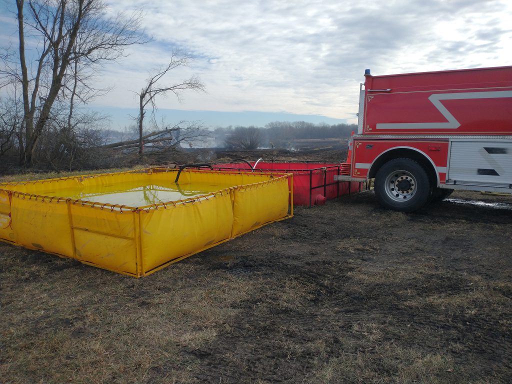 Yellow and red tank filled with water sit next to the back end of a red fire engine in an open field.