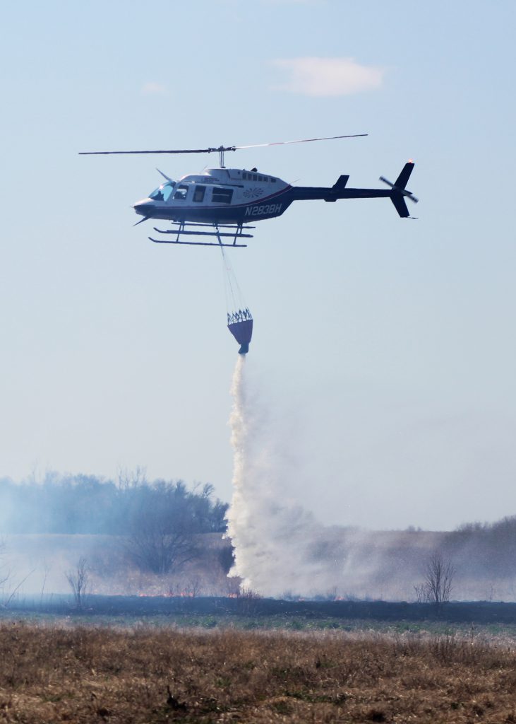 Helicopter drops water from a bucket over a grass field.