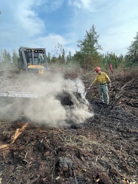 A firefighter uses a hose to spray the hot peat while a dozer in the background prepares to push the peat.