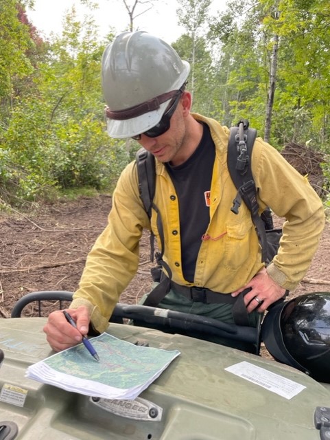 A male firefighter holds a pen in his right hand up to a colored map on top of a UTV in the field.