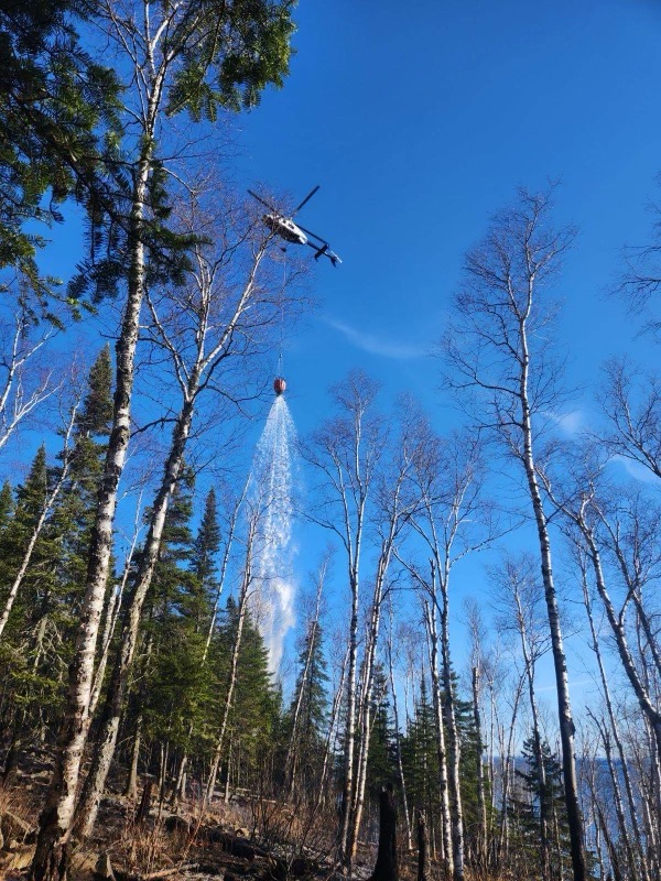 Image of a forest landscape against blue sky with a helicopter dropping water from a bucket. 