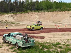 A green and yellow vehicle drive through a gravel pit with trees in the distance.